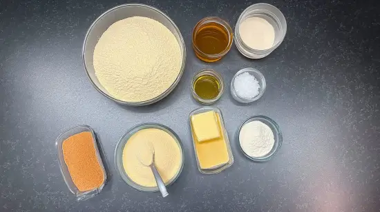 Ingrédients du makrout : semoule, beurre, huile, miel, levure, sel, cannelle, pâte de dattes, farine, eau de fleur d’oranger, tous soigneusement disposés sur un plan de travail noir.