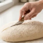 Main utilisant une grignette inclinée pour réaliser le grignage du pain sur un pâton ovale.